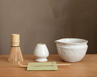 May include: A matcha tea set on a wooden surface. The set includes a bamboo whisk, a small white ceramic vase, a white ceramic bowl, a green cloth, and a bamboo scoop. The background is a solid brown color.