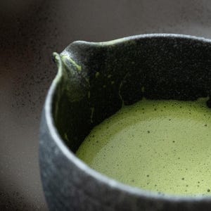 Pu&ograve; includere: Primo piano di una ciotola in ceramica grigio scuro piena di t&egrave; matcha verde brillante. Il t&egrave; ha una consistenza schiumosa e frizzante e la ciotola ha una superficie strutturata. La ciotola ha un beccuccio.