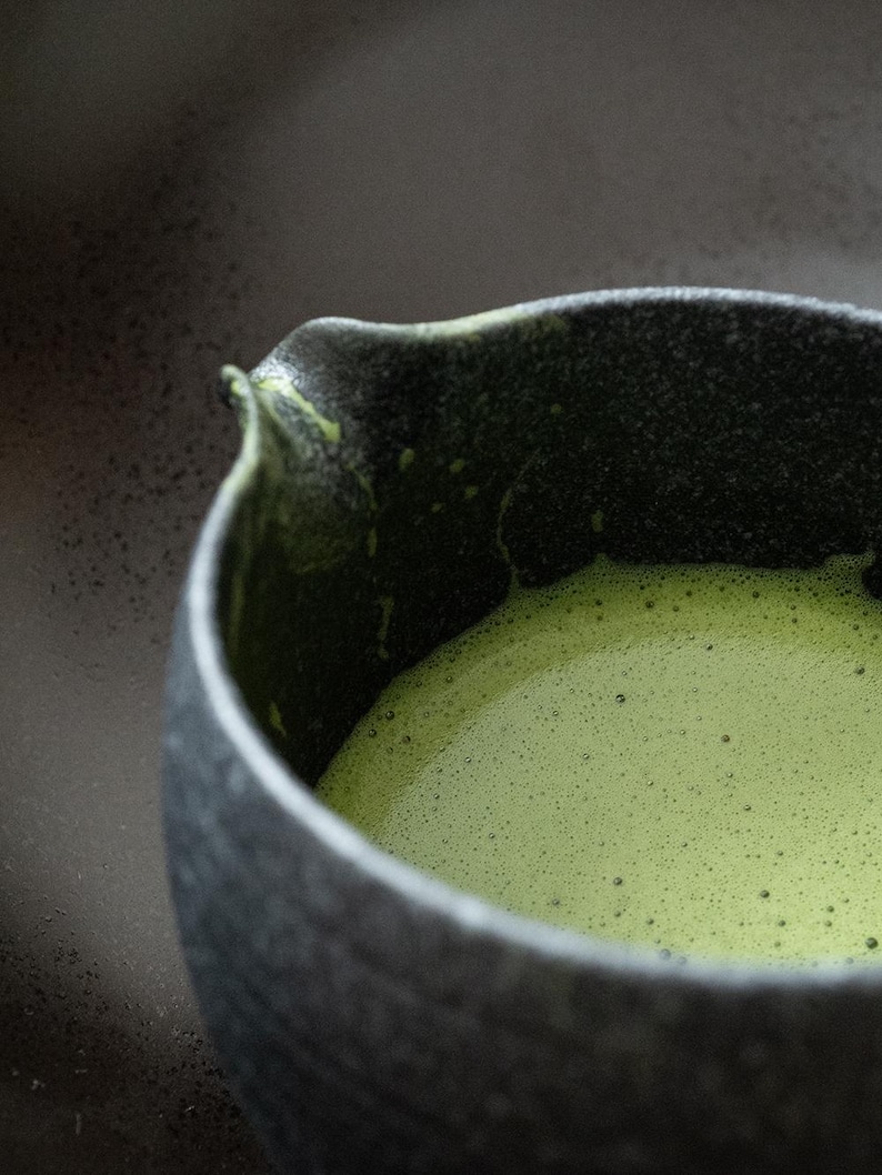 Pu&ograve; includere: Primo piano di una ciotola in ceramica grigio scuro piena di t&egrave; matcha verde brillante. Il t&egrave; ha una consistenza schiumosa e frizzante e la ciotola ha una superficie strutturata. La ciotola ha un beccuccio.