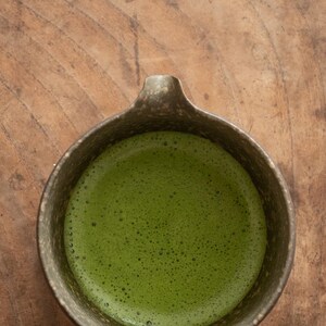 Pu&ograve; includere: Vista dall'alto di una ciotola di ceramica piena di t&egrave; matcha verde brillante. La ciotola ha un esterno scuro e strutturato e un piccolo manico. La superficie del t&egrave; &egrave; ricoperta da minuscole bolle, a indicare che &egrave; appena montato.
