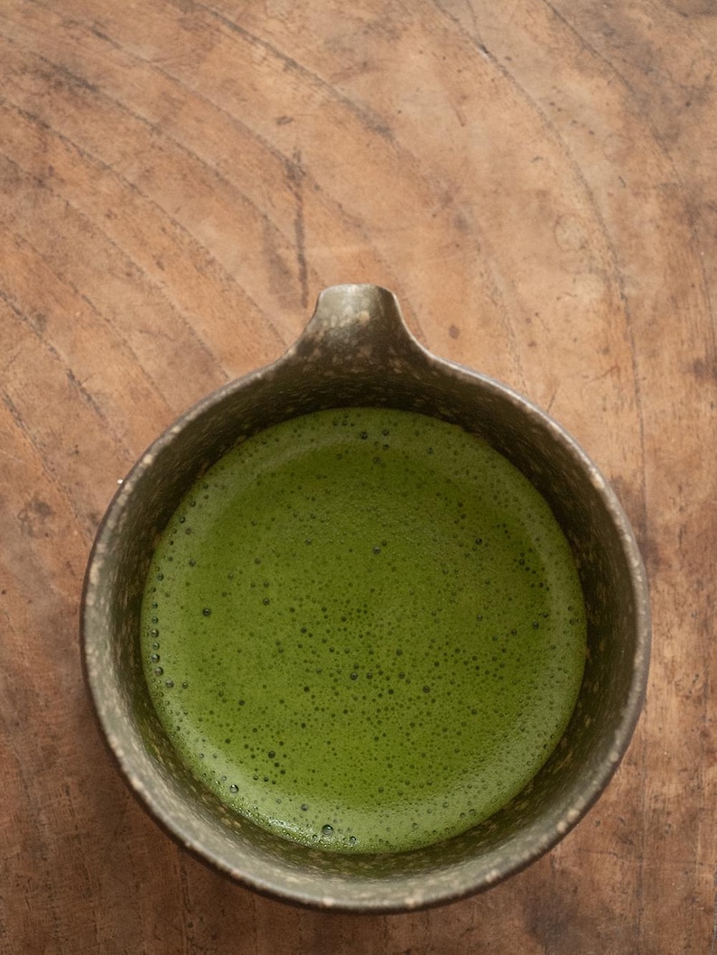 Pu&ograve; includere: Vista dall'alto di una ciotola di ceramica piena di t&egrave; matcha verde brillante. La ciotola ha un esterno scuro e strutturato e un piccolo manico. La superficie del t&egrave; &egrave; ricoperta da minuscole bolle, a indicare che &egrave; appena montato.