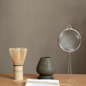 Pu&ograve; includere: Un set da t&egrave; matcha con un frullino di bamb&ugrave;, una ciotola di ceramica, un colino in acciaio inossidabile, un cucchiaio di bamb&ugrave; e un panno bianco. Il frullino &egrave; marrone chiaro, la ciotola &egrave; verde scuro e il colino &egrave; argentato. Il set &egrave; disposto su una superficie di legno.
