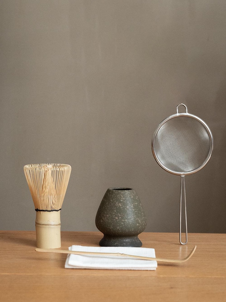 Pu&ograve; includere: Un set da t&egrave; matcha con un frullino di bamb&ugrave;, una ciotola di ceramica, un colino in acciaio inossidabile, un cucchiaio di bamb&ugrave; e un panno bianco. Il frullino &egrave; marrone chiaro, la ciotola &egrave; verde scuro e il colino &egrave; argentato. Il set &egrave; disposto su una superficie di legno.