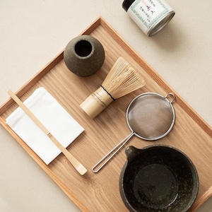 Pu&ograve; includere: Un vassoio di legno contiene un set da t&egrave; matcha. Il set include una ciotola in ceramica, una frusta, un cucchiaio, un setaccio e un piccolo contenitore con testo giapponese. Gli oggetti sono disposti su una superficie chiara.