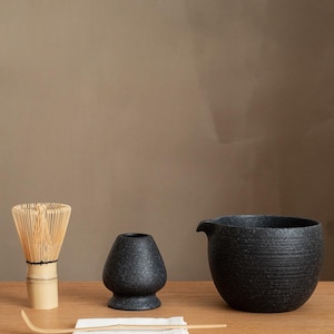 Pu&ograve; includere: Un set da t&egrave; matcha su una superficie di legno. Il set include una frusta di bamb&ugrave;, una ciotola di ceramica nera, un contenitore per il t&egrave; in ceramica nera, un cucchiaio di bamb&ugrave; e un panno bianco.