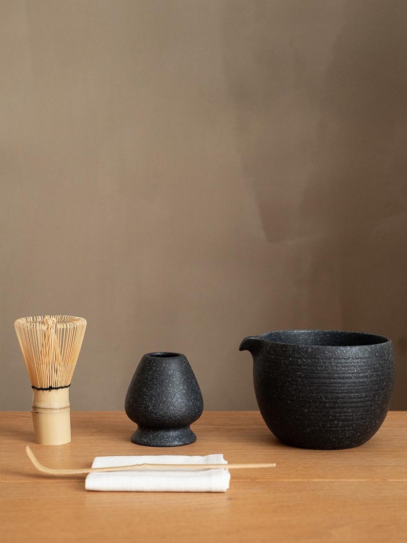 Pu&ograve; includere: Un set da t&egrave; matcha su una superficie di legno. Il set include una frusta di bamb&ugrave;, una ciotola di ceramica nera, un contenitore per il t&egrave; in ceramica nera, un cucchiaio di bamb&ugrave; e un panno bianco.