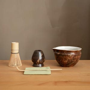 May include: A matcha tea set on a wooden surface. The set includes a bamboo whisk, a small dark brown ceramic tea bowl, a green cloth, a bamboo scoop, and a larger brown ceramic bowl with a white interior.