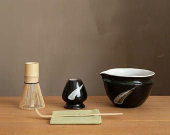 May include: A matcha tea set on a wooden surface. The set includes a bamboo whisk, a black ceramic bowl with a white interior, a black ceramic tea cup, a bamboo scoop, and a green cloth.