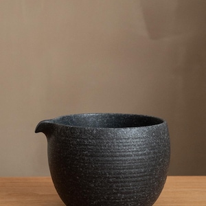 Pu&ograve; includere: Brocca in ceramica nera con superficie strutturata e piccolo beccuccio. La brocca ha un corpo arrotondato e poggia su una superficie di legno. Lo sfondo &egrave; di colore neutro.