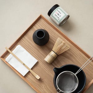 Pu&ograve; includere: Un set da t&egrave; matcha su un vassoio di legno. Il set include una frusta di bamb&ugrave;, una ciotola di ceramica nera, un setaccio di metallo, un piccolo vaso nero, un cucchiaio di bamb&ugrave;, un panno bianco e una lattina di t&egrave; matcha con testo giapponese.