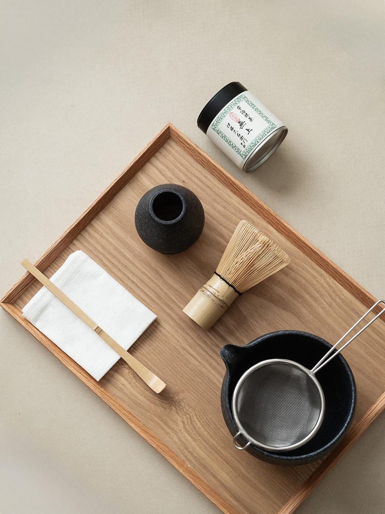 Pu&ograve; includere: Un set da t&egrave; matcha su un vassoio di legno. Il set include una frusta di bamb&ugrave;, una ciotola di ceramica nera, un setaccio di metallo, un piccolo vaso nero, un cucchiaio di bamb&ugrave;, un panno bianco e una lattina di t&egrave; matcha con testo giapponese.