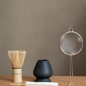 Pu&ograve; includere: Un set da t&egrave; matcha su una superficie di legno. Il set include una frusta di bamb&ugrave;, una ciotola di ceramica scura macchiata, un cucchiaio di bamb&ugrave;, un setaccio in acciaio inossidabile e un panno bianco. Gli articoli sono disposti per la preparazione del t&egrave;.