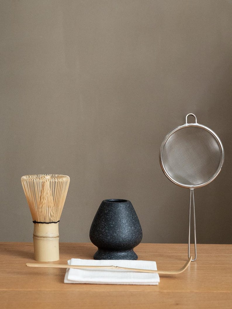 Pu&ograve; includere: Un set da t&egrave; matcha su una superficie di legno. Il set include una frusta di bamb&ugrave;, una ciotola di ceramica scura macchiata, un cucchiaio di bamb&ugrave;, un setaccio in acciaio inossidabile e un panno bianco. Gli articoli sono disposti per la preparazione del t&egrave;.