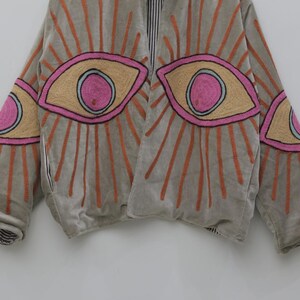May include: A light gray jacket with embroidered eye designs. The eyes are outlined in black, with pink and tan details, and orange sunburst rays. The jacket has a striped lining and a relaxed fit.