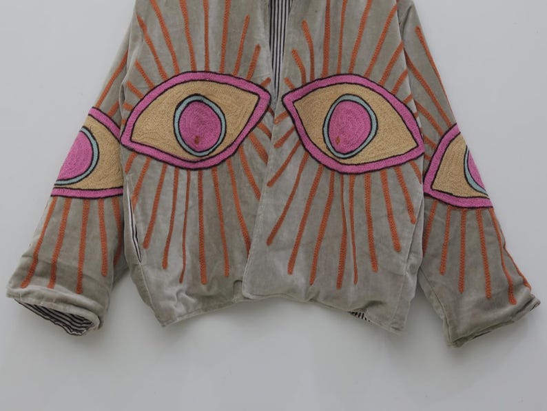 May include: A light gray jacket with embroidered eye designs. The eyes are outlined in black, with pink and tan details, and orange sunburst rays. The jacket has a striped lining and a relaxed fit.