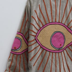 May include: A gray jacket with embroidered eye designs. The jacket features large, stylized eyes on the back and sleeves, with tan, pink, and orange accents. The jacket has a high collar and long sleeves.