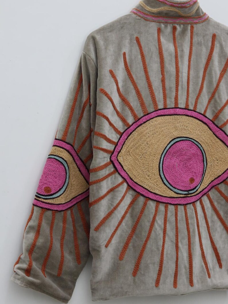 May include: A gray jacket with embroidered eye designs. The jacket features large, stylized eyes on the back and sleeves, with tan, pink, and orange accents. The jacket has a high collar and long sleeves.