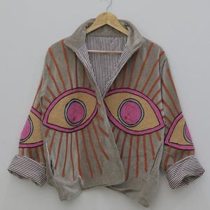 May include: A gray velvet jacket with embroidered eye designs in pink, yellow, and orange. The jacket has a striped lining and cuffs, and is hanging on a wooden hanger against a white background.