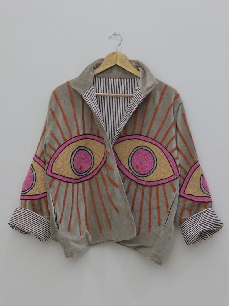 May include: A gray velvet jacket with embroidered eye designs in pink, yellow, and orange. The jacket has a striped lining and cuffs, and is hanging on a wooden hanger against a white background.