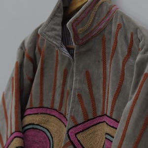 May include: A gray jacket with embroidered sunburst and geometric designs in pink, orange, and tan. The jacket has a high collar and is hanging on a wooden hanger. The lining is striped.