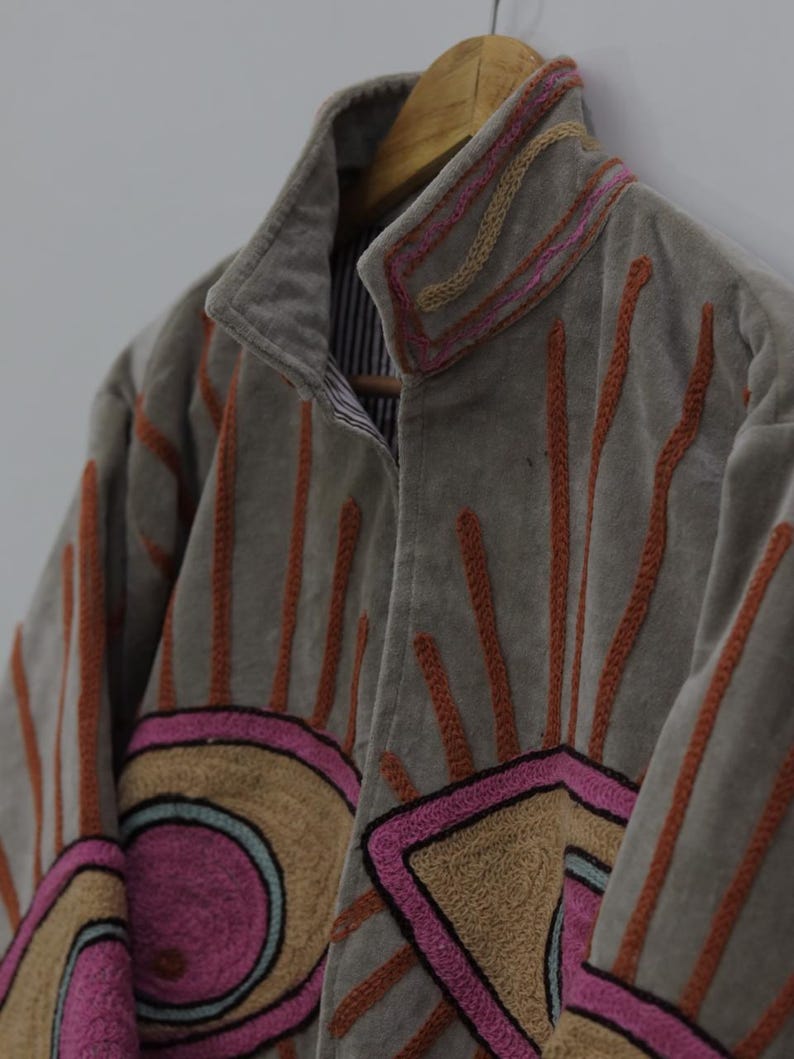 May include: A gray jacket with embroidered sunburst and geometric designs in pink, orange, and tan. The jacket has a high collar and is hanging on a wooden hanger. The lining is striped.