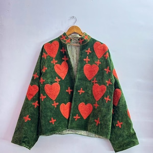 Puede incluir: Una chaqueta de ante verde oscuro con corazones rojos bordados y pequeños símbolos de cruz rojos. La chaqueta tiene cuello alto, mangas largas y un diseño frontal abierto con un forro a rayas de color claro visible en el interior.
