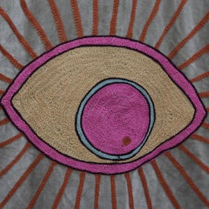 May include: Close-up of a textile with an embroidered eye design. The eye features a pink iris, a light blue outline, and a tan-colored center. The eye is outlined in pink and surrounded by orange rays on a gray background.