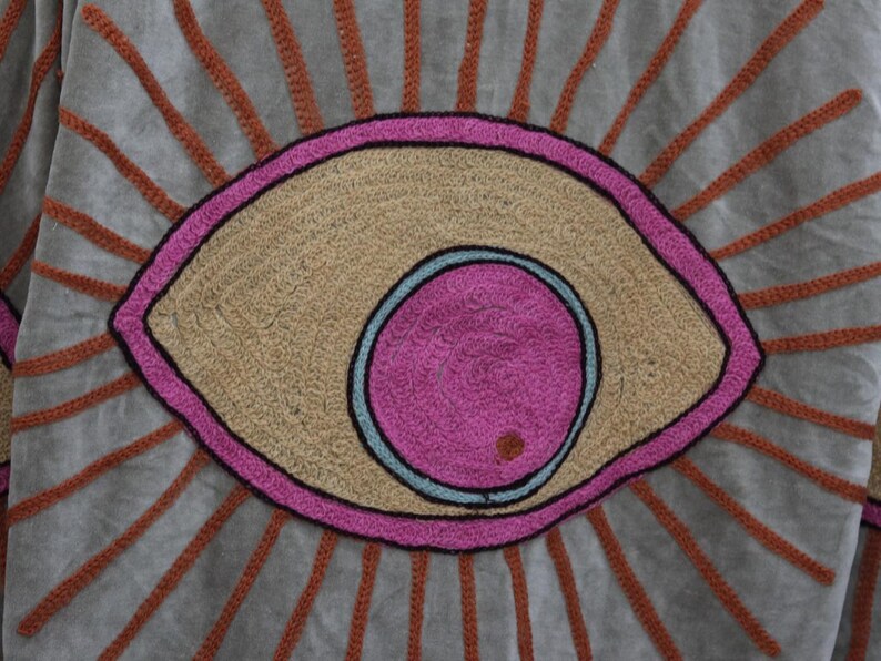 May include: Close-up of a textile with an embroidered eye design. The eye features a pink iris, a light blue outline, and a tan-colored center. The eye is outlined in pink and surrounded by orange rays on a gray background.