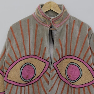 May include: A light gray jacket with embroidered eye designs in pink, tan, and black. The jacket features orange embroidered sunburst patterns and a striped interior. The collar and cuffs have pink and tan embroidery.