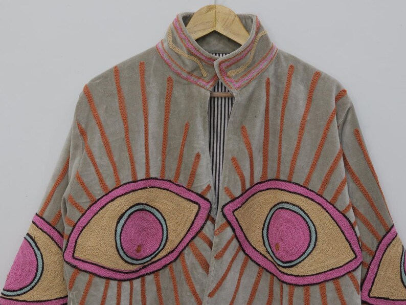 May include: A light gray jacket with embroidered eye designs in pink, tan, and black. The jacket features orange embroidered sunburst patterns and a striped interior. The collar and cuffs have pink and tan embroidery.
