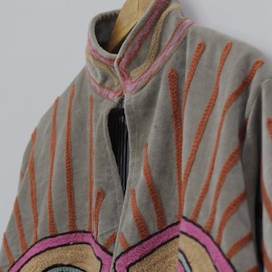 May include: A gray jacket with orange, pink, and tan embroidered details. The jacket has a high collar and is hanging on a wooden hanger. The design includes vertical lines and geometric shapes.