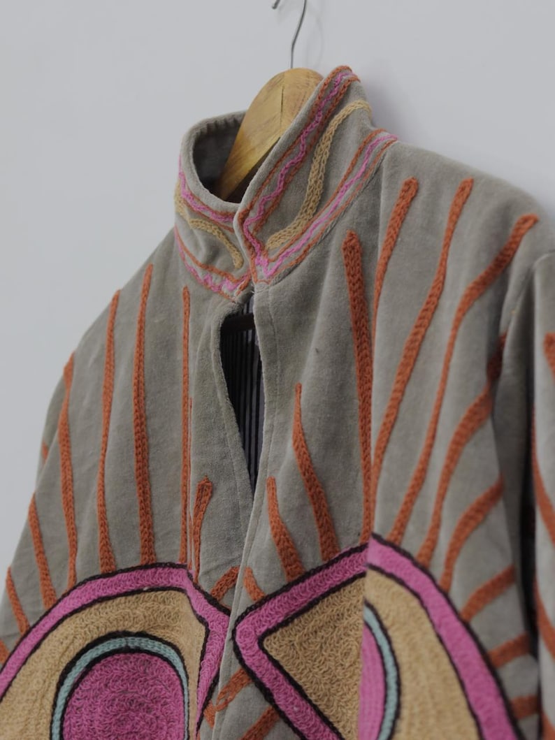 May include: A gray jacket with orange, pink, and tan embroidered details. The jacket has a high collar and is hanging on a wooden hanger. The design includes vertical lines and geometric shapes.