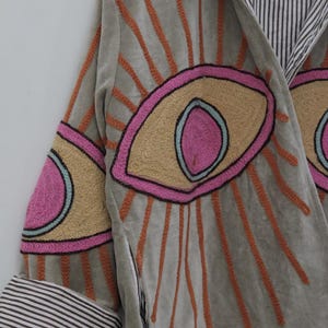 May include: A light gray jacket with embroidered eye designs in pink, tan, and orange. The jacket features a striped lining and cuffs. The sleeves have eye designs with orange rays emanating from them.