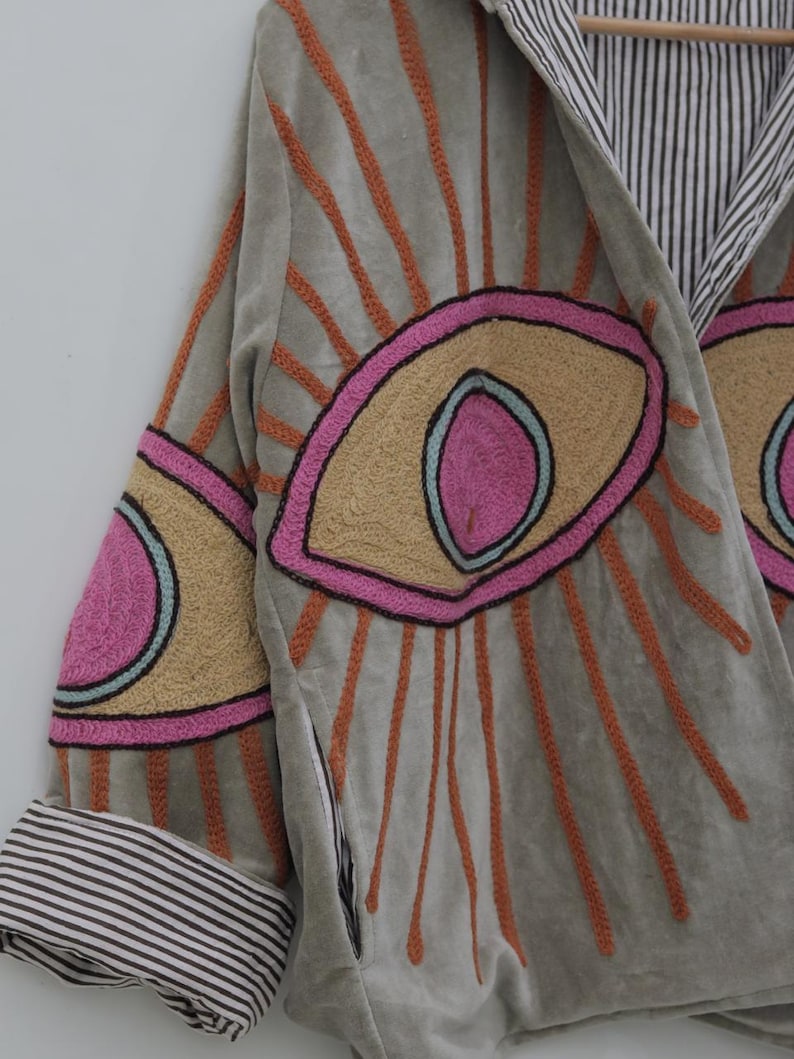 May include: A light gray jacket with embroidered eye designs in pink, tan, and orange. The jacket features a striped lining and cuffs. The sleeves have eye designs with orange rays emanating from them.