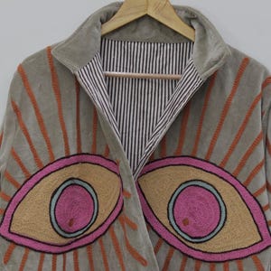 May include: A gray velvet jacket with embroidered eye designs. The jacket features orange sunburst patterns and large eye motifs in pink, tan, and blue. The interior lining has a black and white striped pattern.