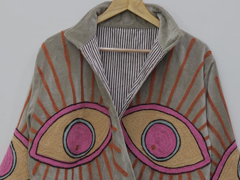May include: A gray velvet jacket with embroidered eye designs. The jacket features orange sunburst patterns and large eye motifs in pink, tan, and blue. The interior lining has a black and white striped pattern.