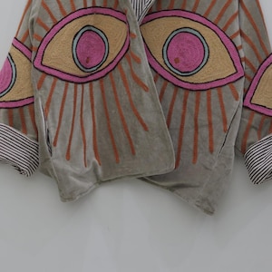May include: A gray jacket with embroidered eye designs in pink, tan, and orange. The jacket has striped cuffs and a draped front. The eye designs are surrounded by orange rays.