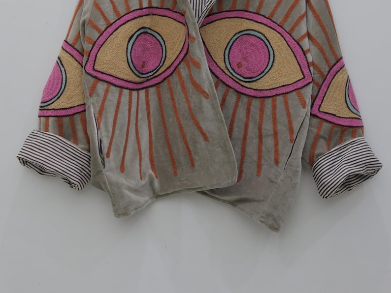 May include: A gray jacket with embroidered eye designs in pink, tan, and orange. The jacket has striped cuffs and a draped front. The eye designs are surrounded by orange rays.
