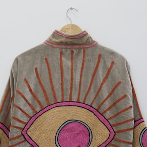 May include: A tan jacket with a high collar and embroidered sunburst design. The sunburst features orange rays and a pink and tan eye-like shape. The jacket is hanging on a wooden hanger.