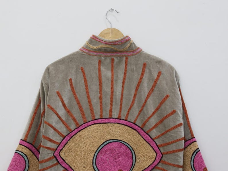 May include: A tan jacket with a high collar and embroidered sunburst design. The sunburst features orange rays and a pink and tan eye-like shape. The jacket is hanging on a wooden hanger.