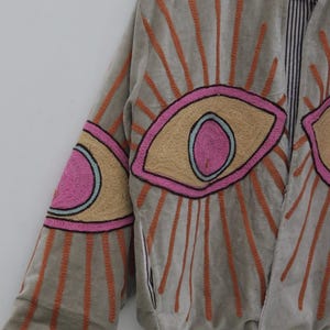 May include: A gray jacket with embroidered eye designs. The jacket features large, stylized eyes in pink, tan, and blue, with orange sunburst patterns radiating from them. The jacket's sleeves and collar are also detailed with embroidery.