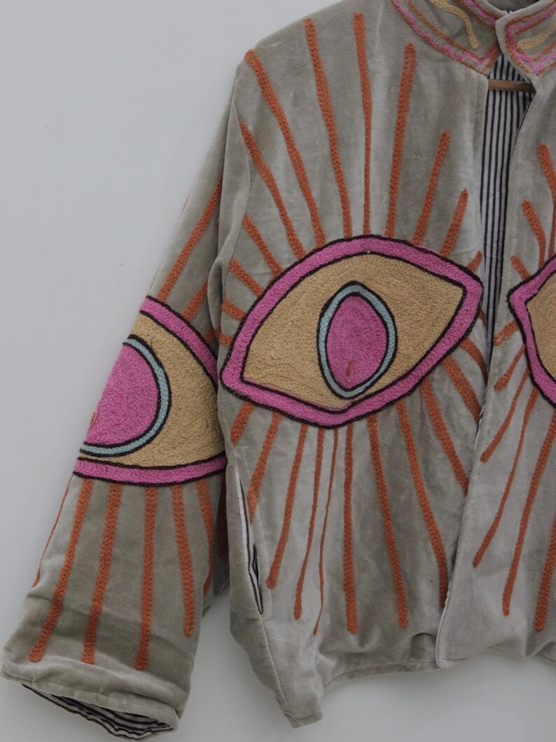May include: A gray jacket with embroidered eye designs. The jacket features large, stylized eyes in pink, tan, and blue, with orange sunburst patterns radiating from them. The jacket's sleeves and collar are also detailed with embroidery.