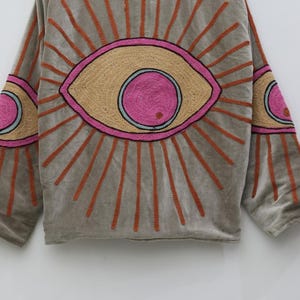 May include: A gray, long-sleeved top with embroidered eye designs. The eyes are in shades of pink, tan, and orange, with orange sunburst rays emanating from them. The sleeves also feature eye designs.