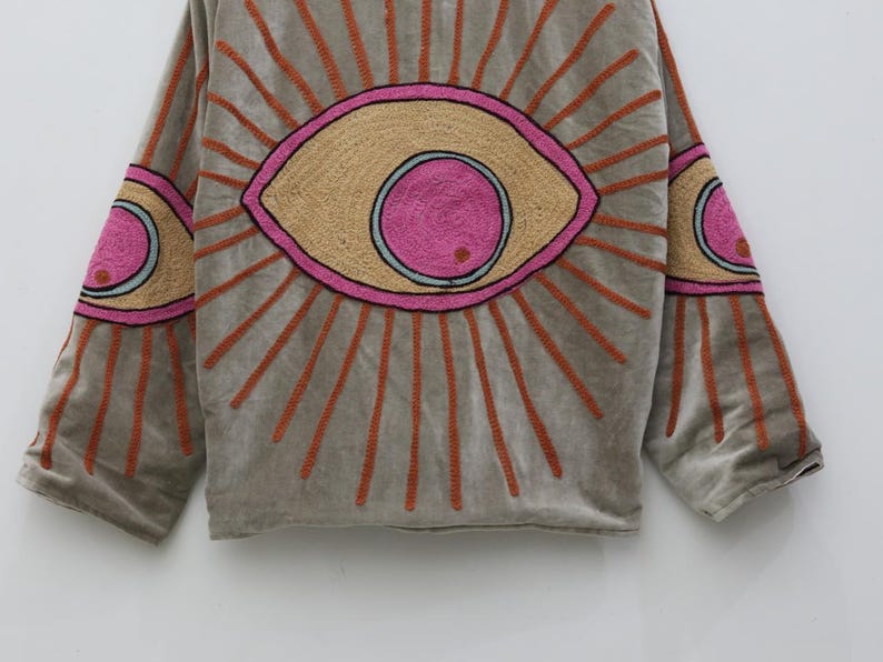 May include: A gray, long-sleeved top with embroidered eye designs. The eyes are in shades of pink, tan, and orange, with orange sunburst rays emanating from them. The sleeves also feature eye designs.