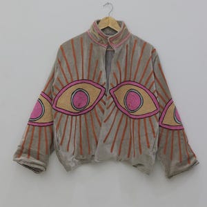 May include: A gray jacket with embroidered eye designs in pink, tan, and orange. The jacket has a high collar and is hanging on a wooden hanger. The lining is striped.