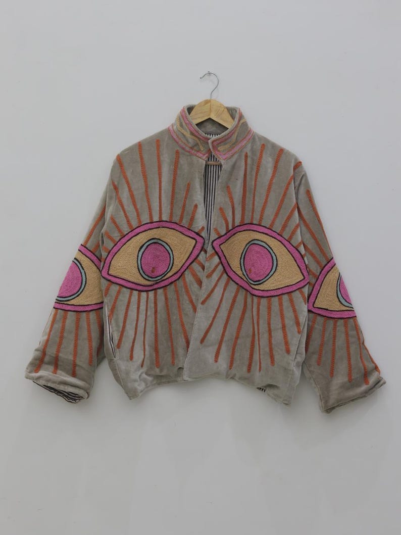 May include: A gray jacket with embroidered eye designs in pink, tan, and orange. The jacket has a high collar and is hanging on a wooden hanger. The lining is striped.