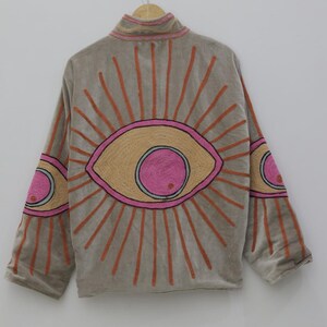 May include: A gray jacket with a high collar and long sleeves, featuring an embroidered eye design on the back and sleeves. The eye design includes pink, orange, and tan colors, with orange rays emanating from the eye.