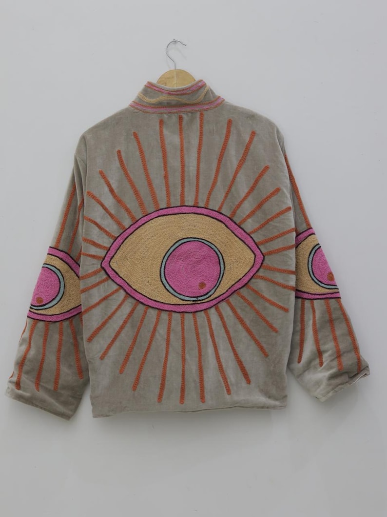 May include: A gray jacket with a high collar and long sleeves, featuring an embroidered eye design on the back and sleeves. The eye design includes pink, orange, and tan colors, with orange rays emanating from the eye.
