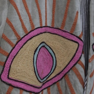 May include: Close-up of a light green jacket with an embroidered eye design. The eye is outlined in pink and black, with a tan center and a blue inner detail. Orange embroidered rays emanate from the eye, creating a sunburst effect.