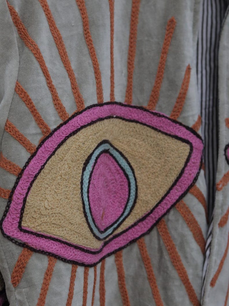 May include: Close-up of a light green jacket with an embroidered eye design. The eye is outlined in pink and black, with a tan center and a blue inner detail. Orange embroidered rays emanate from the eye, creating a sunburst effect.
